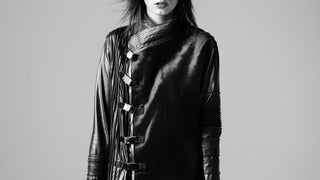 Black and white photo of a person wearing a long black coat with leather sleeves on a plain background