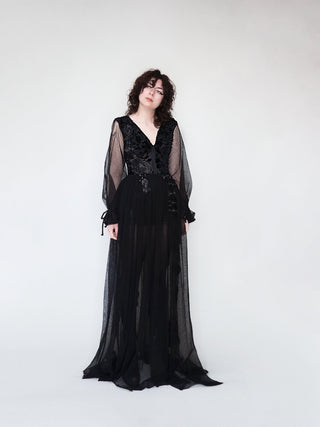A person standing against a plain background, wearing a long, black, silk gown with French lace appliques and a corseted midriff.
