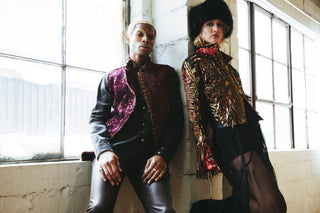 Two people in stylish outfits leaning against a wall with large windows.