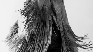 Person wearing a fringed jacket with a plain background carrying a fringed purse.
