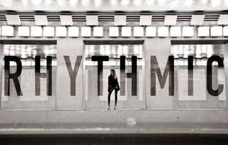 Person standing in front of a large word 'RHYTHMIC' on a wall.